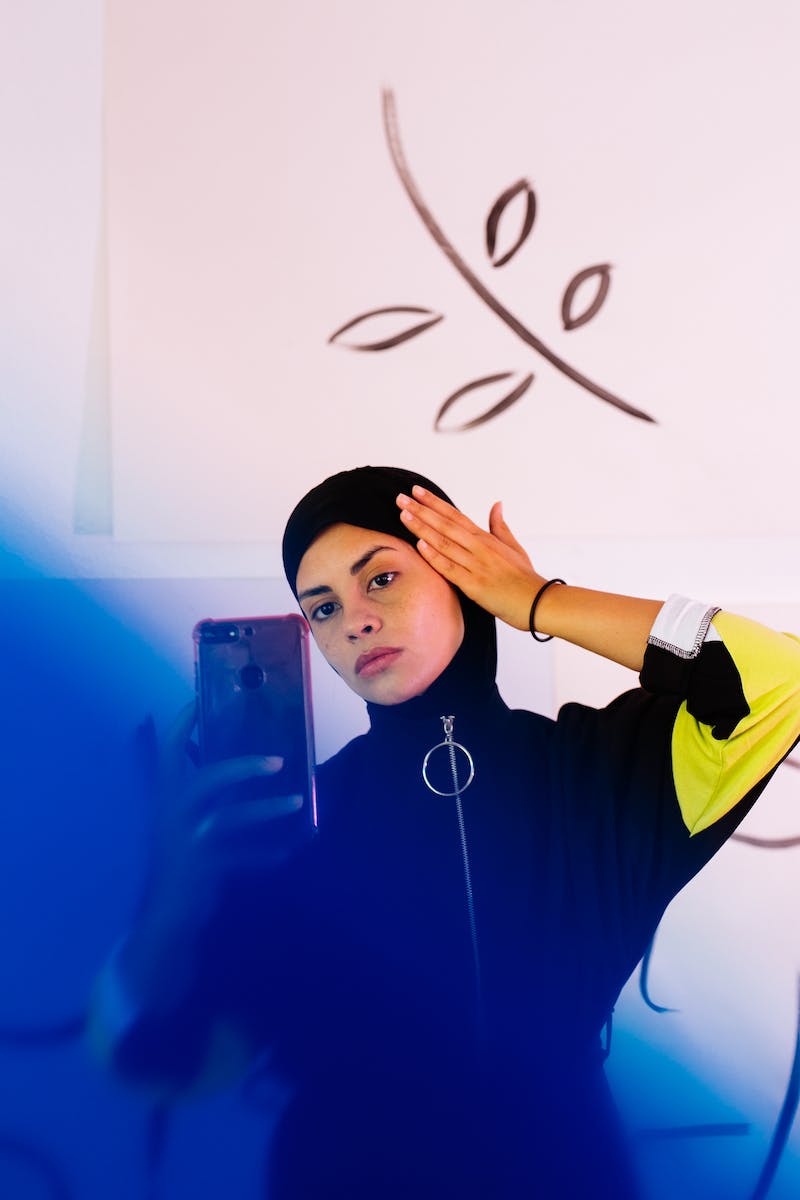 Concentrated young ethnic female wearing sportswear with tight hood covering head and taking selfie against white wall with abstract illustrations
