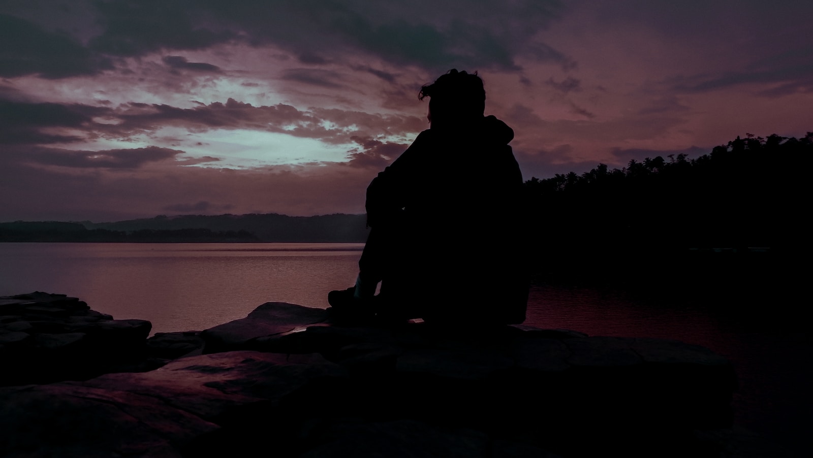 silhouette of man sitting on rock near body of water during sunset